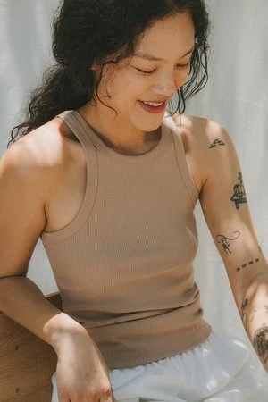 Smiling young Asian woman in ribbed beige tank top with arm tattoos, sitting indoors with natural light, looking downwards.