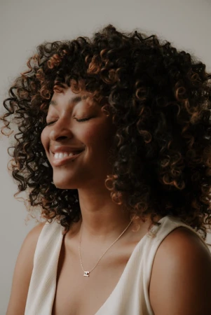 Close-up portrait of a smiling Black woman with voluminous curly hair wearing a white top and a delicate necklace against a plain wall.