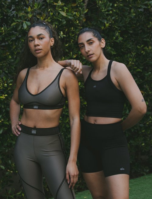 Two women in athletic wear posing outdoors against a green foliage backdrop, showcasing activewear fashion and fitness lifestyle.