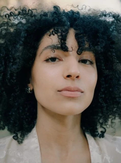 Close-up portrait of an adult woman with dark curly hair, fair skin, and neutral expression, wearing a pale patterned robe indoors.