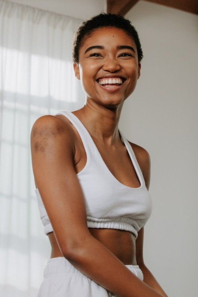 Smiling Black woman with short hair wearing white athletic top against a bright indoor background by Kris Ryan
