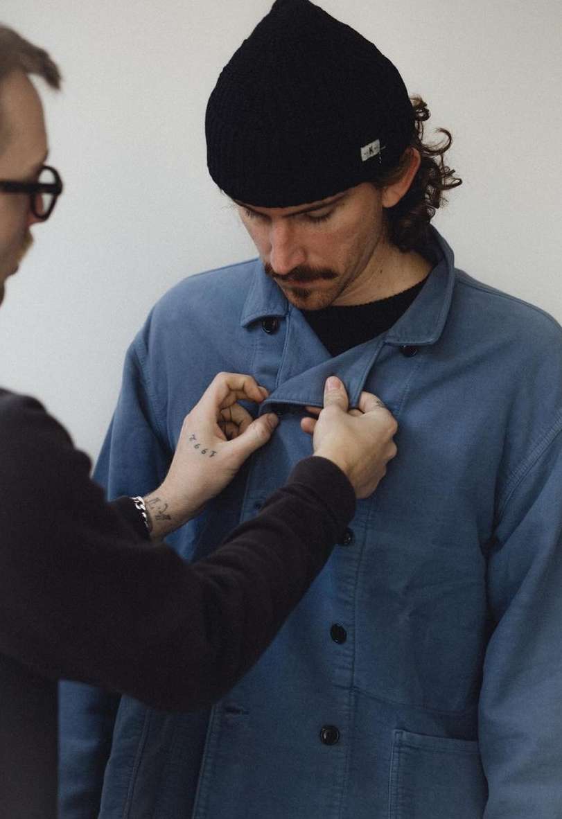 Man wearing a blue work jacket being buttoned by another person in a bright setting, showcasing fashion and styling.