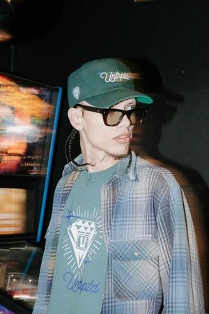 Young adult wearing a baseball cap and flannel shirt stands indoors near a vintage arcade game, illuminated with colorful neon lights.
