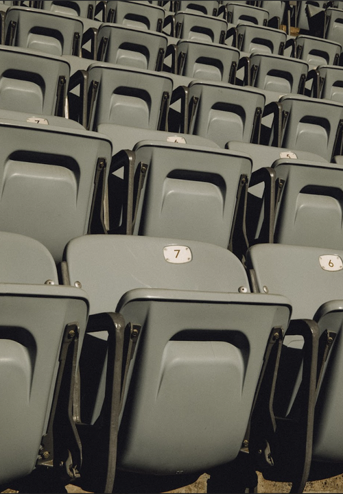 Rows of empty gray seats, numbered 6 and 7, are visible in an outdoor venue, with a simple geometric design and a muted color palette.
