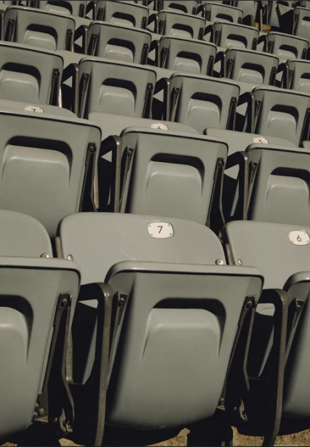 Rows of empty gray seats, numbered 6 and 7, are visible in an outdoor venue, with a simple geometric design and a muted color palette.