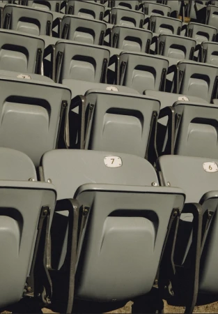 Rows of empty gray seats, numbered 6 and 7, are visible in an outdoor venue, with a simple geometric design and a muted color palette.
