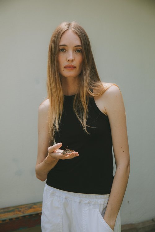 Young woman stands outdoors holding a butterfly in her hand. She wears a black top and white pants, with a plain wall behind her.