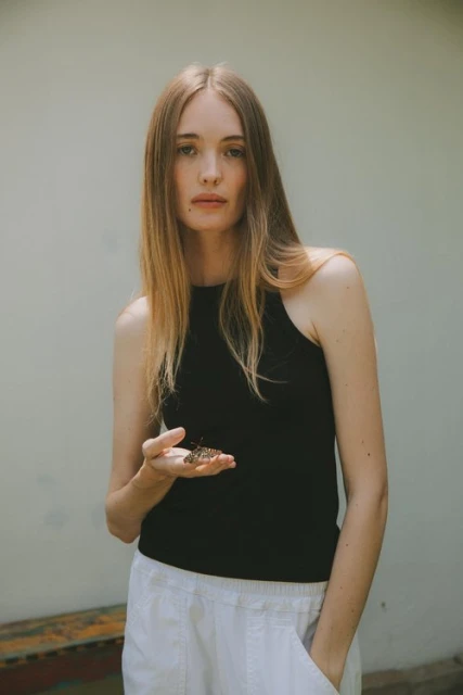 Young woman stands outdoors holding a butterfly in her hand. She wears a black top and white pants, with a plain wall behind her.