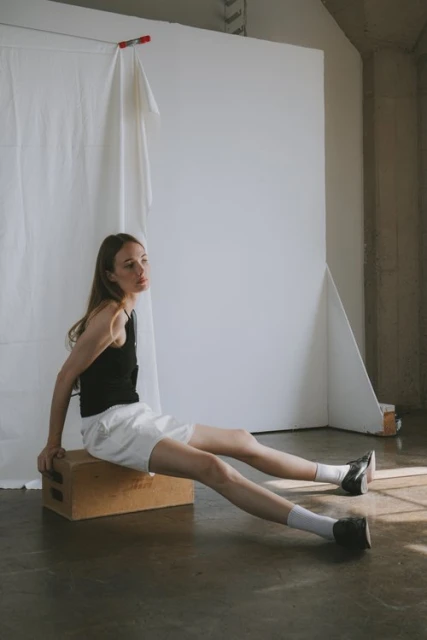 Woman in black top and white shorts sitting on wood box in bright indoor studio, wearing socks and loafers, looking off-camera.
