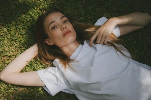 Woman lying on the grass in white top, outdoors, in natural lighting. She is resting with her hands behind her head.