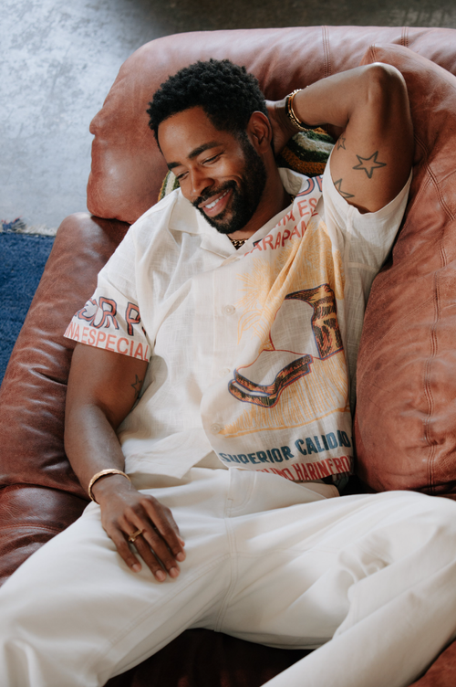 Man in printed shirt lounges on leather couch smiling, wearing white pants and jewelry with a star tattoo on his arm.
