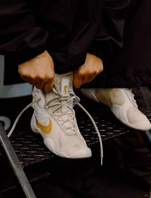A close-up of a person tying the laces of white and gold Nike wrestling shoes while sitting on metal stairs in an indoor setting.
