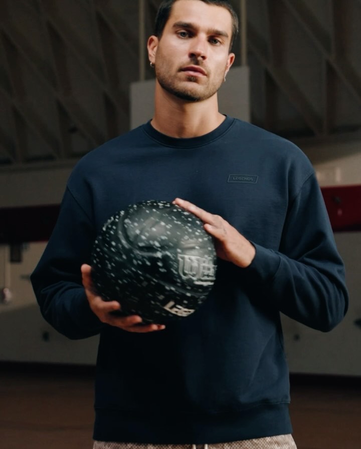 Athletic man in a blue sweatshirt holding a Wilson basketball indoors, posing in a gymnasium with natural light.