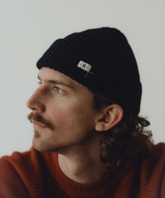 Headshot of a young adult man wearing a black knit beanie and rust-colored sweater, posing indoors against a neutral background. 