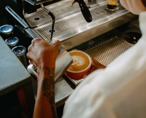 Close-up of a barista pouring steamed milk into a red ceramic coffee cup with latte art, next to an espresso machine.