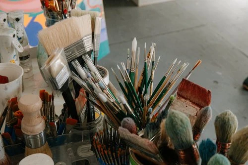 Close-up studio shot of various paintbrushes in holders and jars, arranged on a white table in an artist's studio, painting in background.