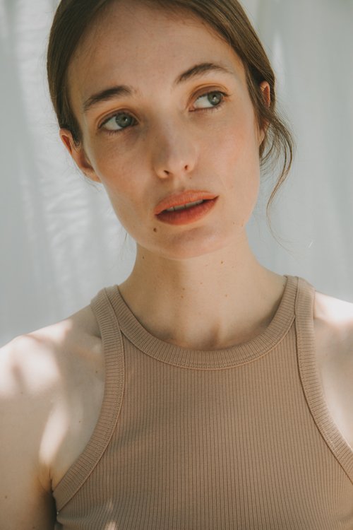Close-up studio portrait of a fair-skinned adult woman with blue eyes, wearing a beige tank top, looking upwards in soft light.