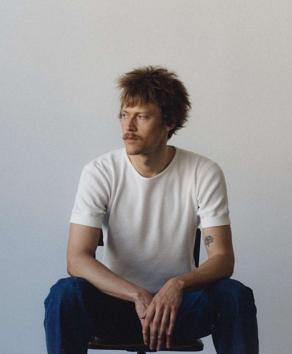 Portrait of a man with a mustache sitting on a chair against a white background, photographed by Kris Ryan.