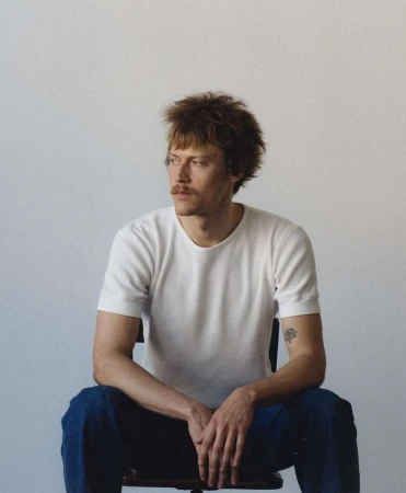 Portrait of a man with a mustache sitting on a chair against a white background, photographed by Kris Ryan.