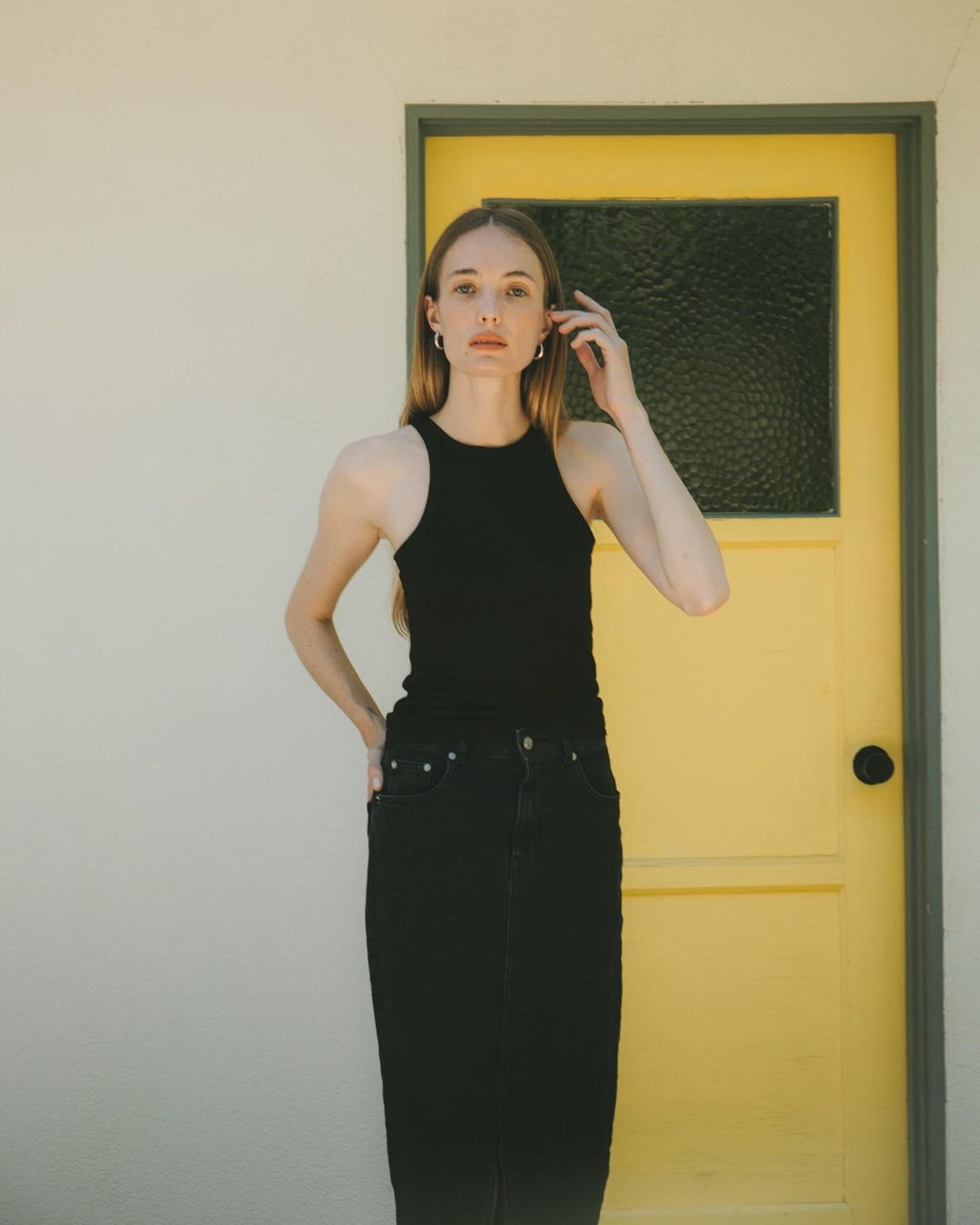 Blonde woman in a black top and skirt posing in front of a yellow door. Neutral background, natural lighting, studio setting. Fashion photography.