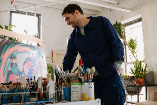 Smiling artist in a blue jacket stands in an art studio with his hand in his pocket looking at an abstract painting with paintbrushes.