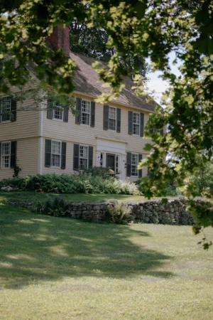 Exterior of a colonial-style house featuring a light-yellow facade, dark shutters, multi-paned windows, and a lush green lawn.