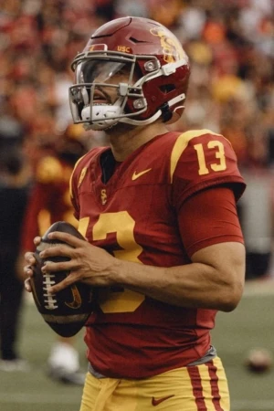 American football player wearing a helmet and jersey number 13 holding the football in a stadium with spectators in the background.