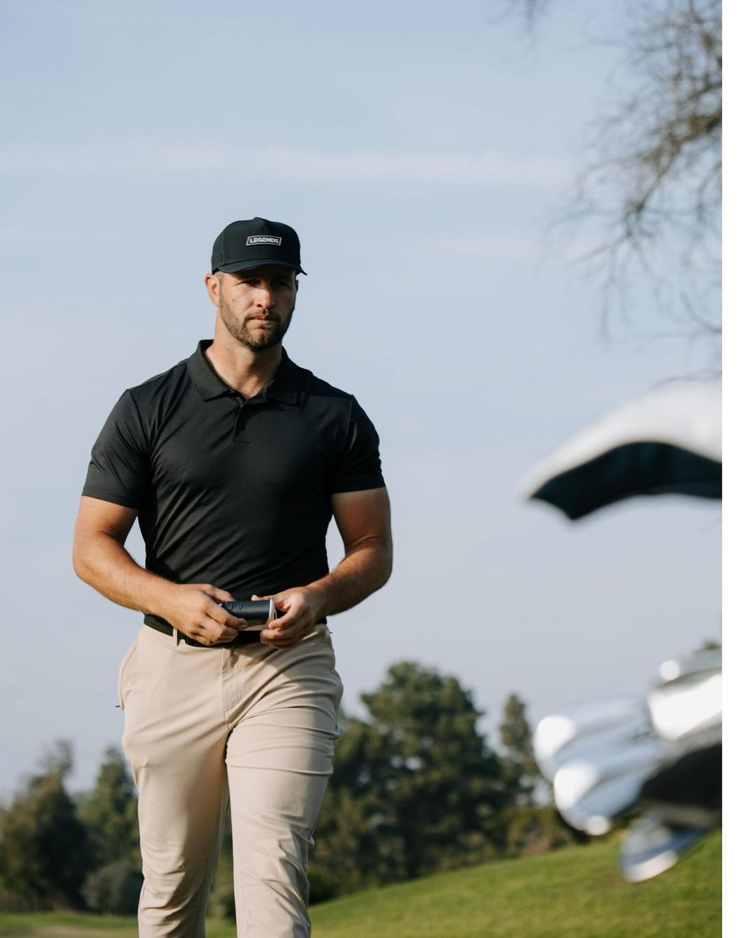 Man in black polo and khaki pants on green grass with golf clubs. Golfing attire and outdoor sport create a sense of activity.