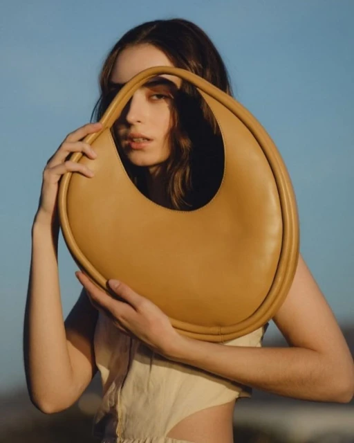 Woman holds an tan hobo-style purse up to her face against a clear blue sky. She wears a beige top, and her brown hair is wavy.