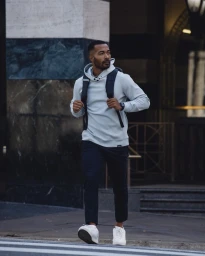 Black man in a light grey hoodie and dark trousers walking across a city street with a dark backpack.