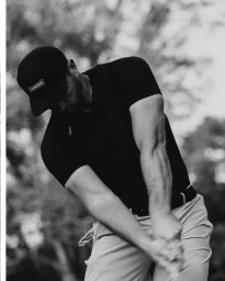 Man wearing a black baseball cap and polo shirt swinging a golf club outdoors.