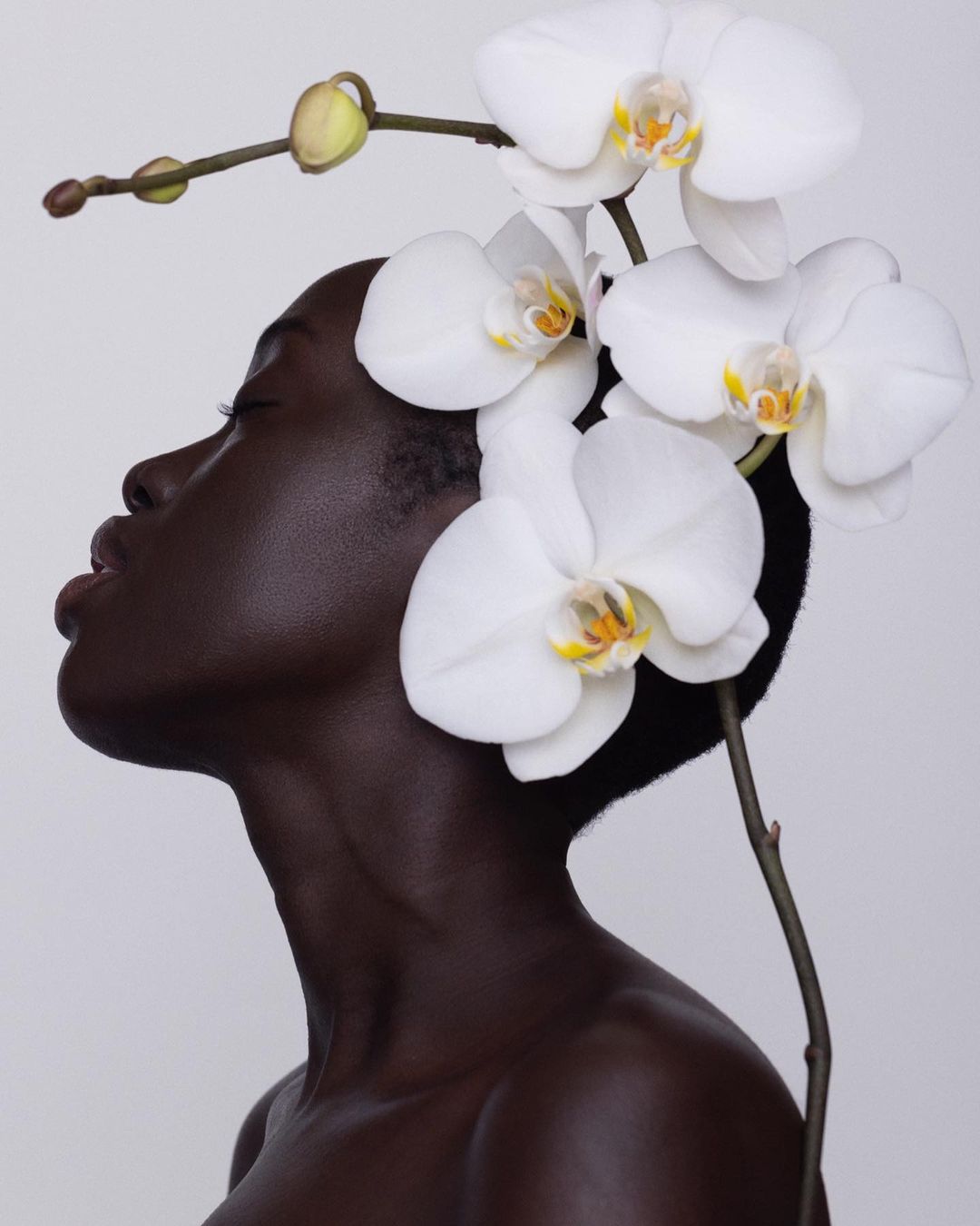 Close-up of a dark-skinned woman with closed eyes and an orchid flower headpiece against a white background in a beauty editorial shoot.