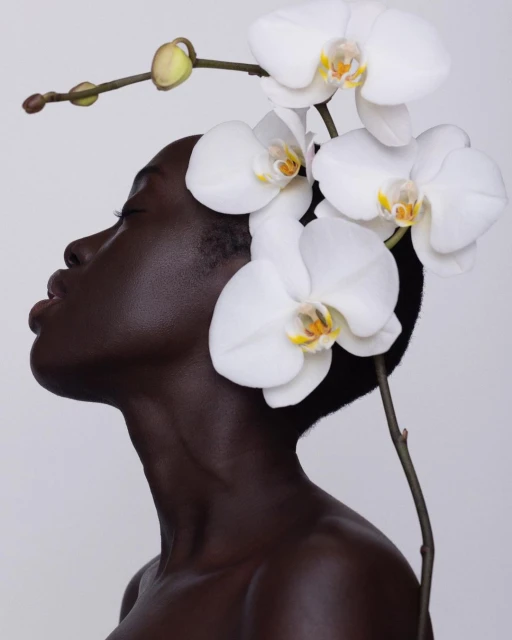 Close-up of a dark-skinned woman with closed eyes and an orchid flower headpiece against a white background in a beauty editorial shoot.