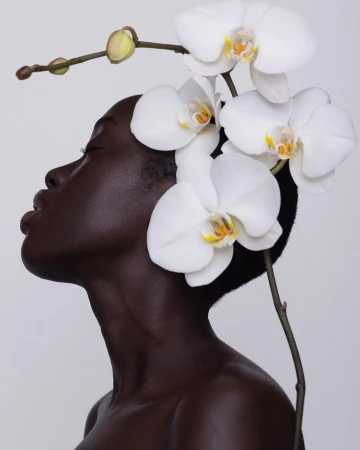 Close-up of a dark-skinned woman with closed eyes and an orchid flower headpiece against a white background in a beauty editorial shoot.