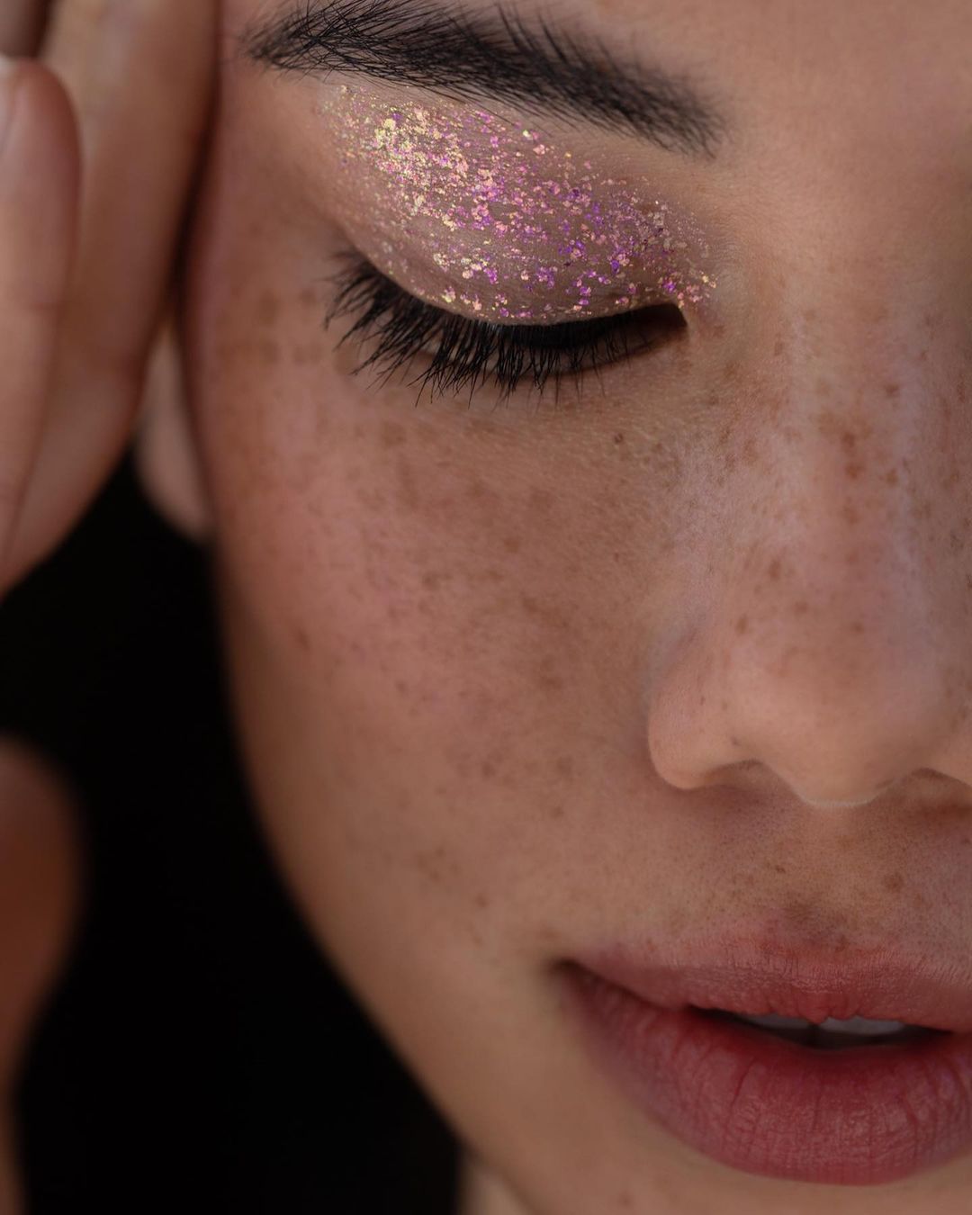 Close-up beauty portrait of a woman with freckles wearing sparkling iridescent eye shadow and placing her hands near her face