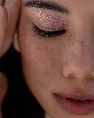 Close-up beauty portrait of a woman with freckles wearing sparkling iridescent eye shadow and placing her hands near her face