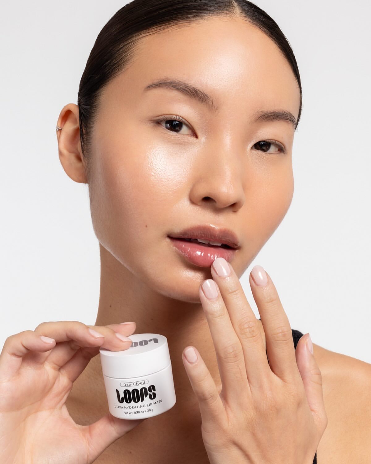 Close-up portrait of an East Asian woman holding a small white LOOPS jar and touching her glossy lips.