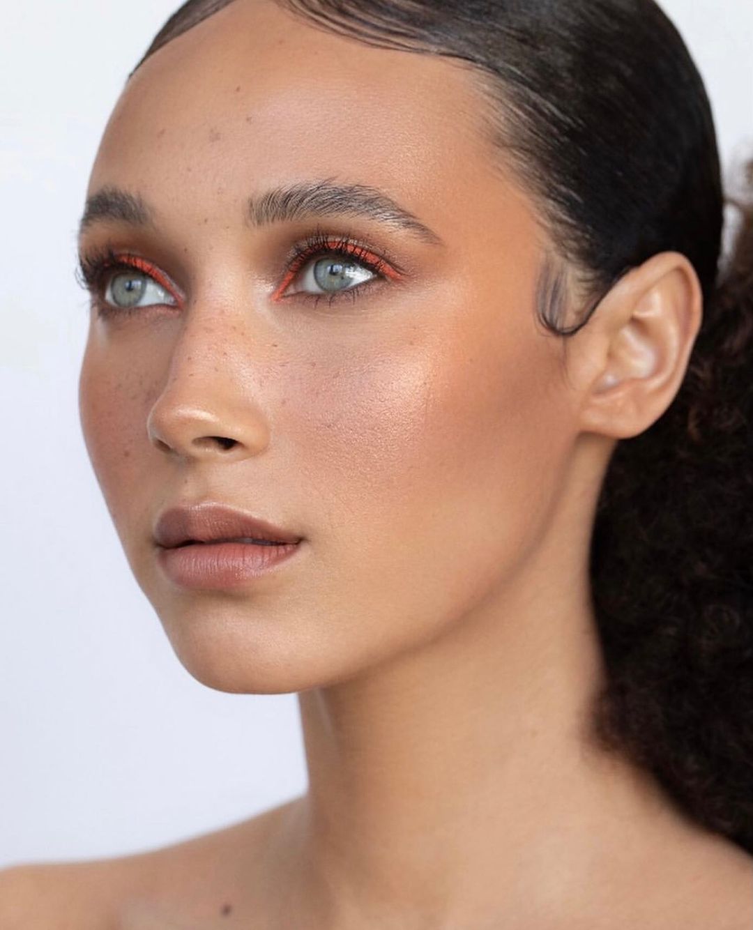 Close-up portrait of a woman with fair skin and freckles wearing copper eyeliner and nude lipstick against a light background.
