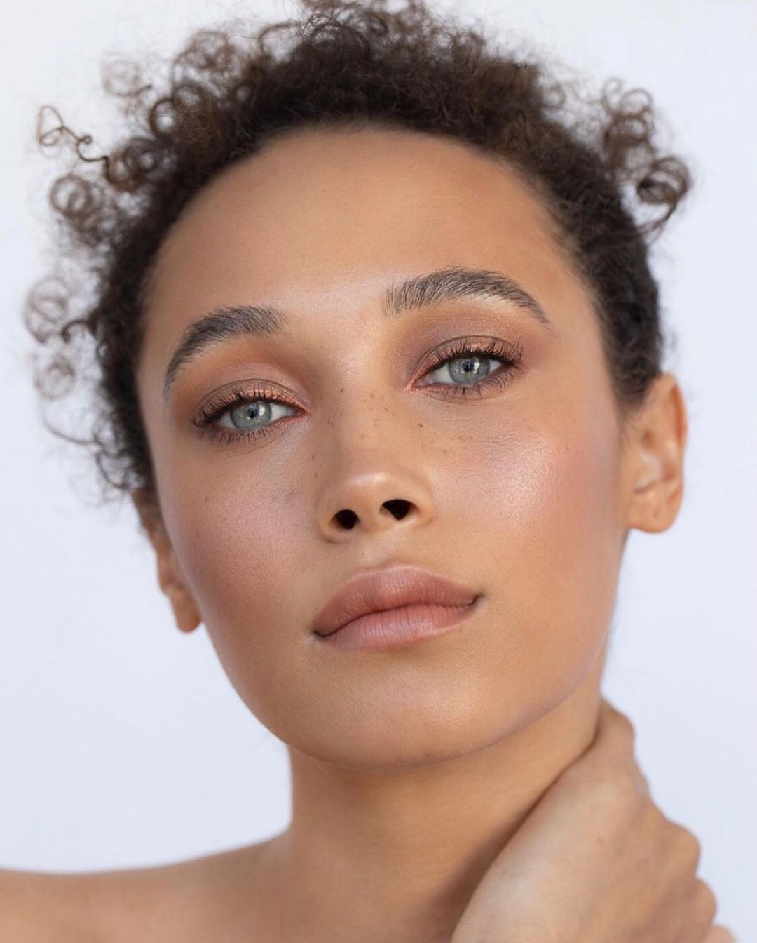 Close-up portrait of a woman with curly brown hair, light eyes, and neutral makeup against a plain white background.
