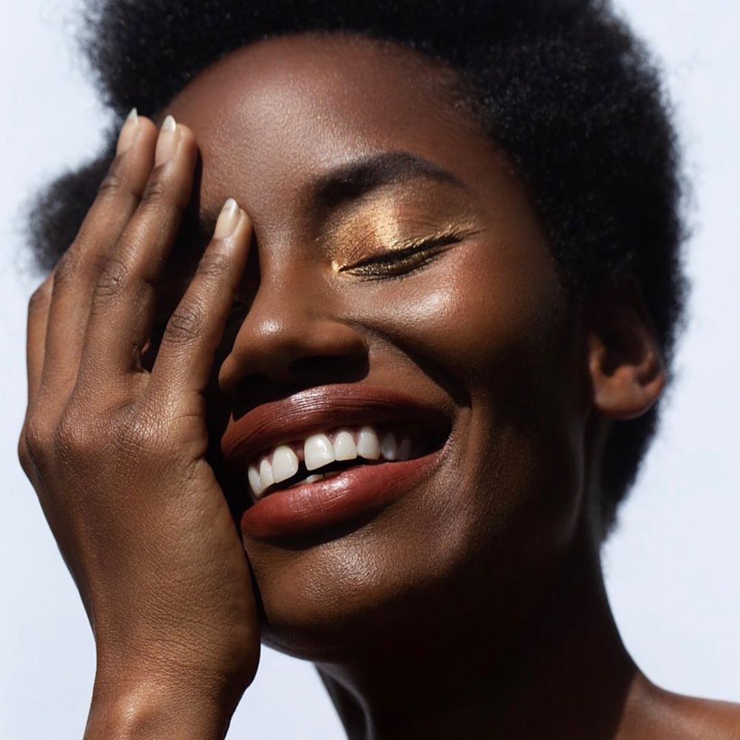 Smiling young African American woman with hand near her face, wearing gold eyeshadow in a studio environment with bright lighting.