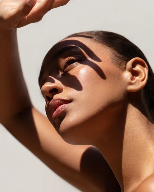 Close-up studio shot of a dark-skinned adult female model shielding her face from the sun, with strong shadows creating visual interest.