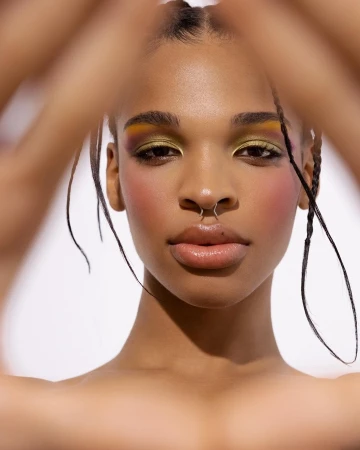 Close-up portrait of a Black woman with colorful eye makeup, braided hair, and a septum ring against a plain white background.