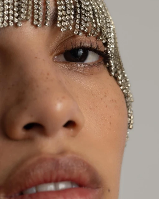 Close-up of a woman's face with her eye, nose, and mouth partially visible, adorned with a jeweled headpiece with hanging strands.