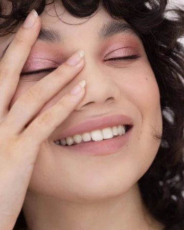 Close-up beauty portrait of a smiling woman with glossy pink eyeshadow and soft matte lips, partially covering her face with her hand.
