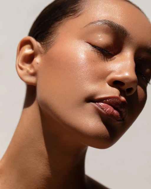 Close-up portrait of a young adult woman with fair skin, shiny lips, and wet-look hair, illuminated by soft, diffused natural light.