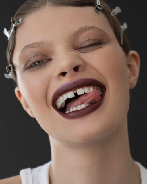 Close-up studio shot of a young adult woman with auburn lipstick playfully sticking out her tongue, against a dark gray background.