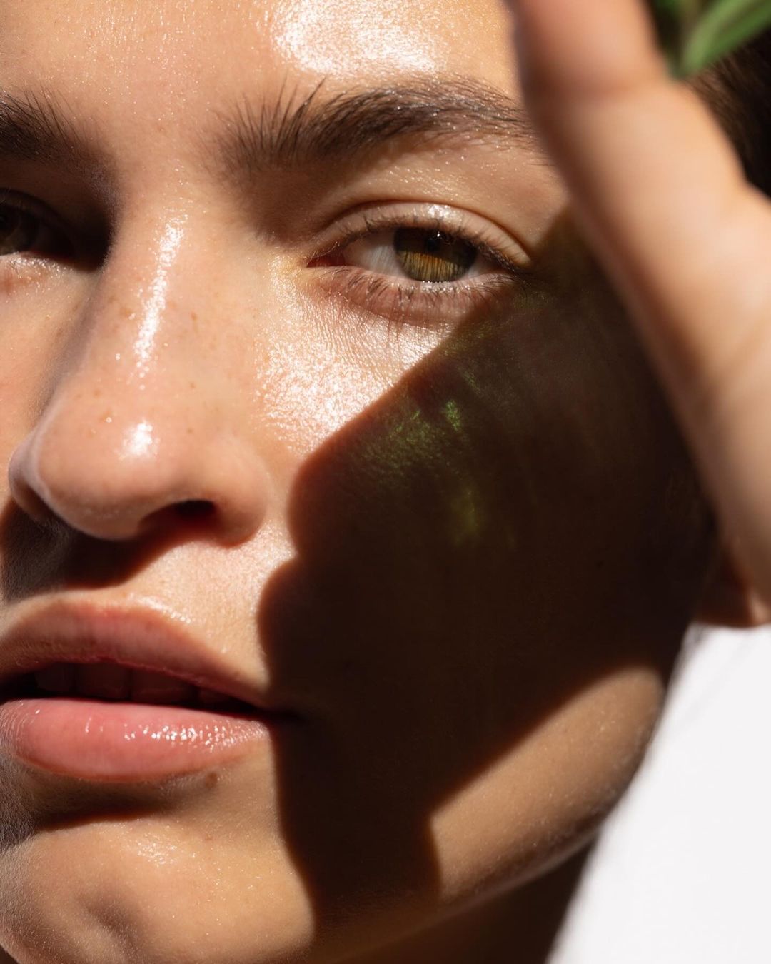 Close-up portrait of a young woman with radiant skin, her hand casting a shadow on her face in natural sunlight, showcasing beauty.