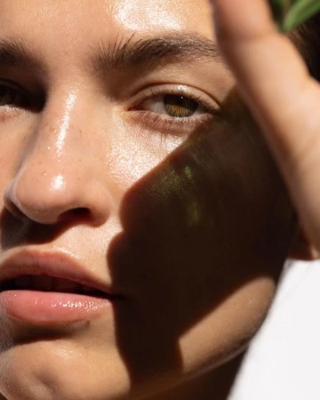 Close-up portrait of a young woman with radiant skin, her hand casting a shadow on her face in natural sunlight, showcasing beauty.