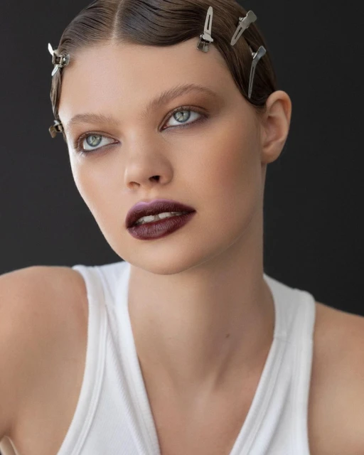 Close-up portrait of a young woman with dark lipstick, styled hair with clips, wearing a white tank top against a dark background.