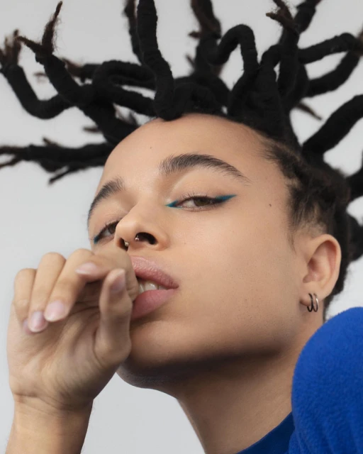 Close-up portrait of a young adult woman with styled dark hair, blue eyeliner, a nose ring, and a blue top against a white backdrop.